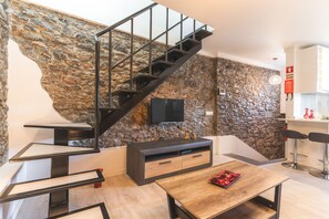 Ein moderner Wohnraum mit Steinwand, einem Holzesstisch, einem an der Wand montierten Fernseher und einer Treppe mit Metallgeländern.