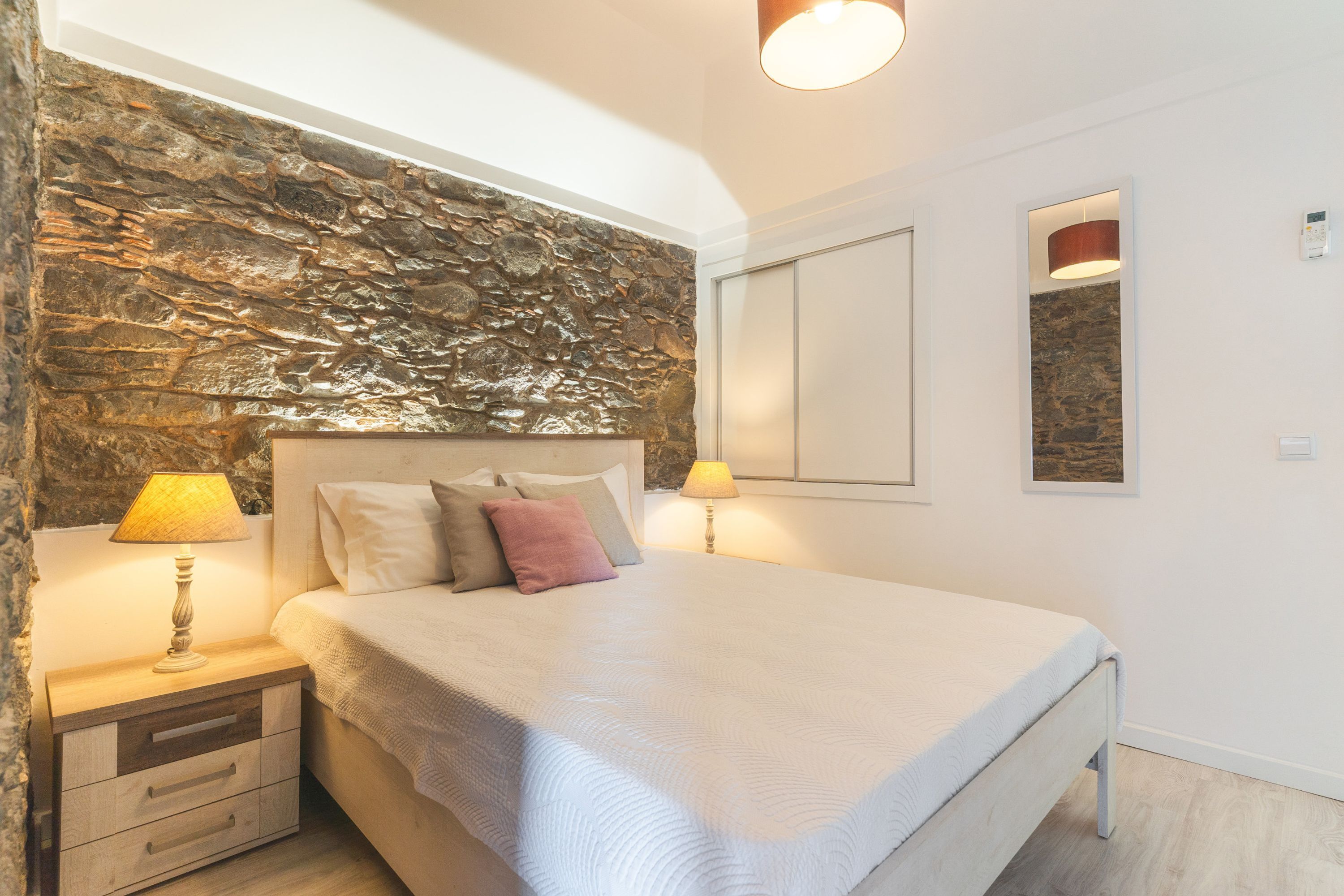 A bedroom with a stone wall, a bed with a white bedspread, two bedside lamps, and a nightstand with drawers.