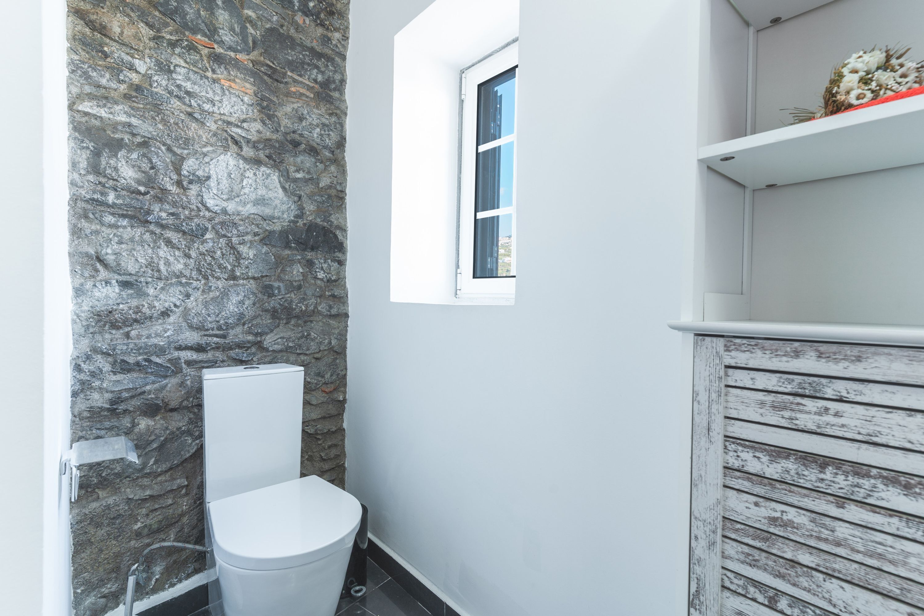 Een badkamer met een stenen muur, een wit toilet, een raam en een witte plank met een decoratief object.