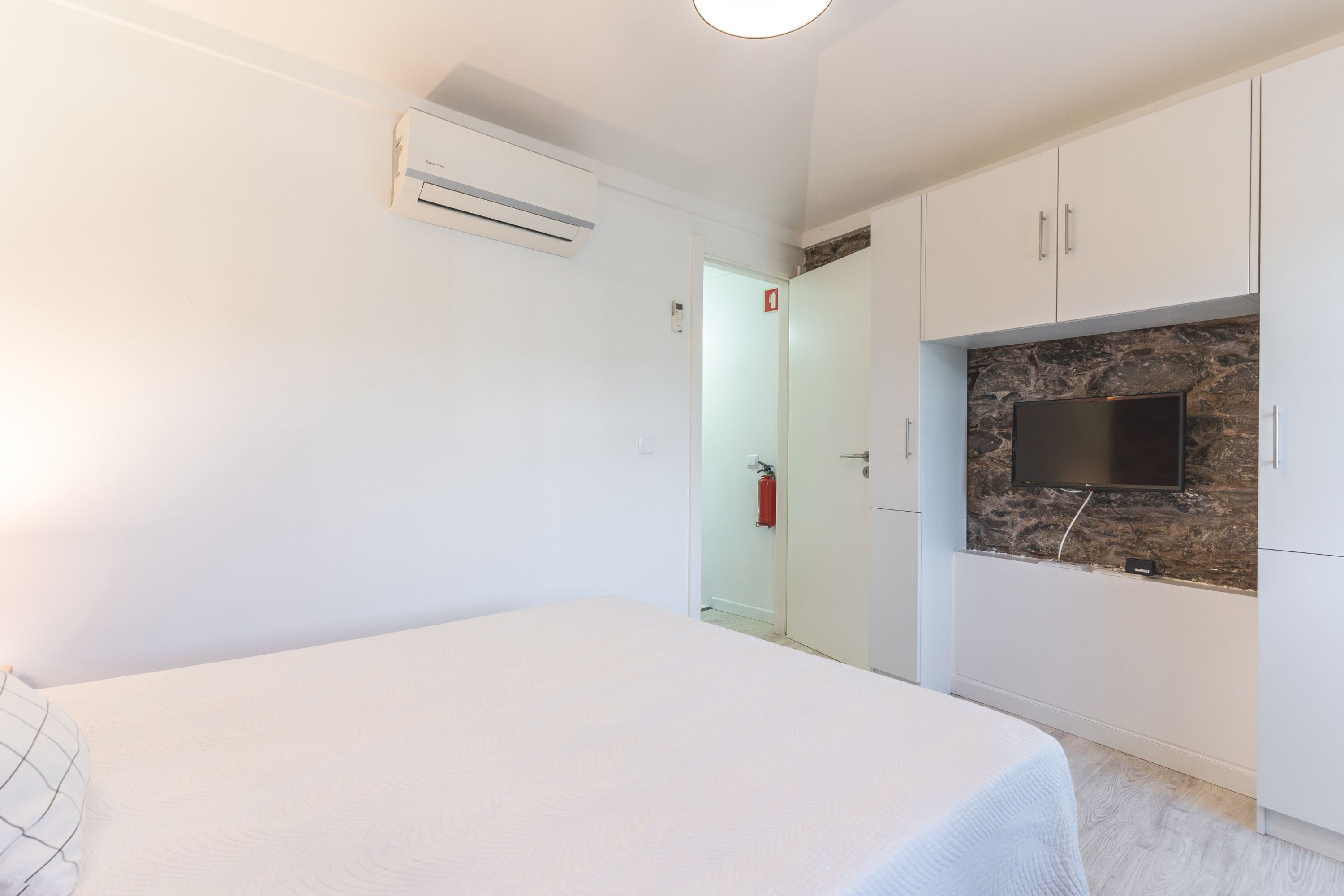 A bedroom with a bed, a TV mounted on a stone wall, and a white wardrobe.