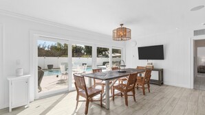 A spacious dining area with a large table and chairs, a television mounted on the wall, and a sliding glass door leading to an outdoor pool area. - Modern Coastal Home in West Palm Beach with Pool (West Palm Beach)