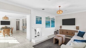 A modern living room with a white brick wall, a flat-screen TV, a dining area with a wooden table and chairs, and a cozy seating area with a sofa and ottoman. - Modern Coastal Home in West Palm Beach with Pool (West Palm Beach)