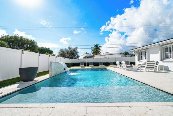 Una piscina rectangular con una cascada, rodeada por una cerca blanca y un edificio blanco con un balcón.