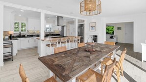 A spacious kitchen with white cabinetry, a large wooden dining table, and a hanging light fixture. - Modern Coastal Home in West Palm Beach with Pool (West Palm Beach)