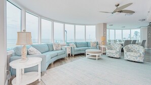 Amplia sala de estar con techo curvo, ventanas amplias que ofrecen vista al mar, un ventilador de techo y una mesa de centro redonda.