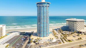 Un hotel alto y cilíndrico con una parte superior circular, situado cerca de una playa de aguas azul cristalino y un estacionamiento al frente.