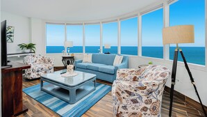 Una sala de estar con un amplio ventanal con vista al océano, un sofá azul, un sillón estampado, una mesa de centro y una lámpara de pie.
