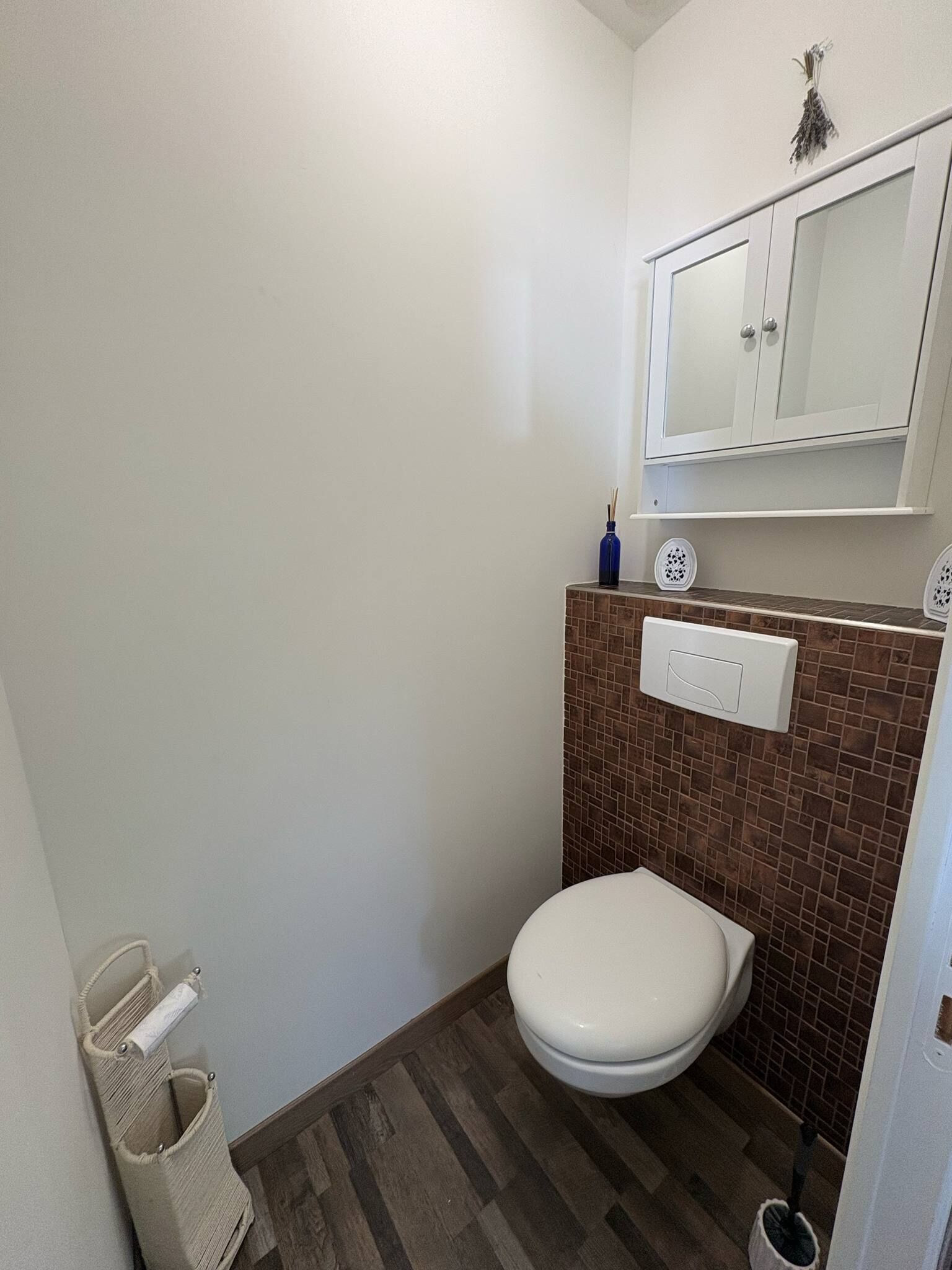 Ein kleines Badezimmer mit Toilette, braunen Fliesen, einem weißen Schrank über der Toilette und einem beigen Boden.
