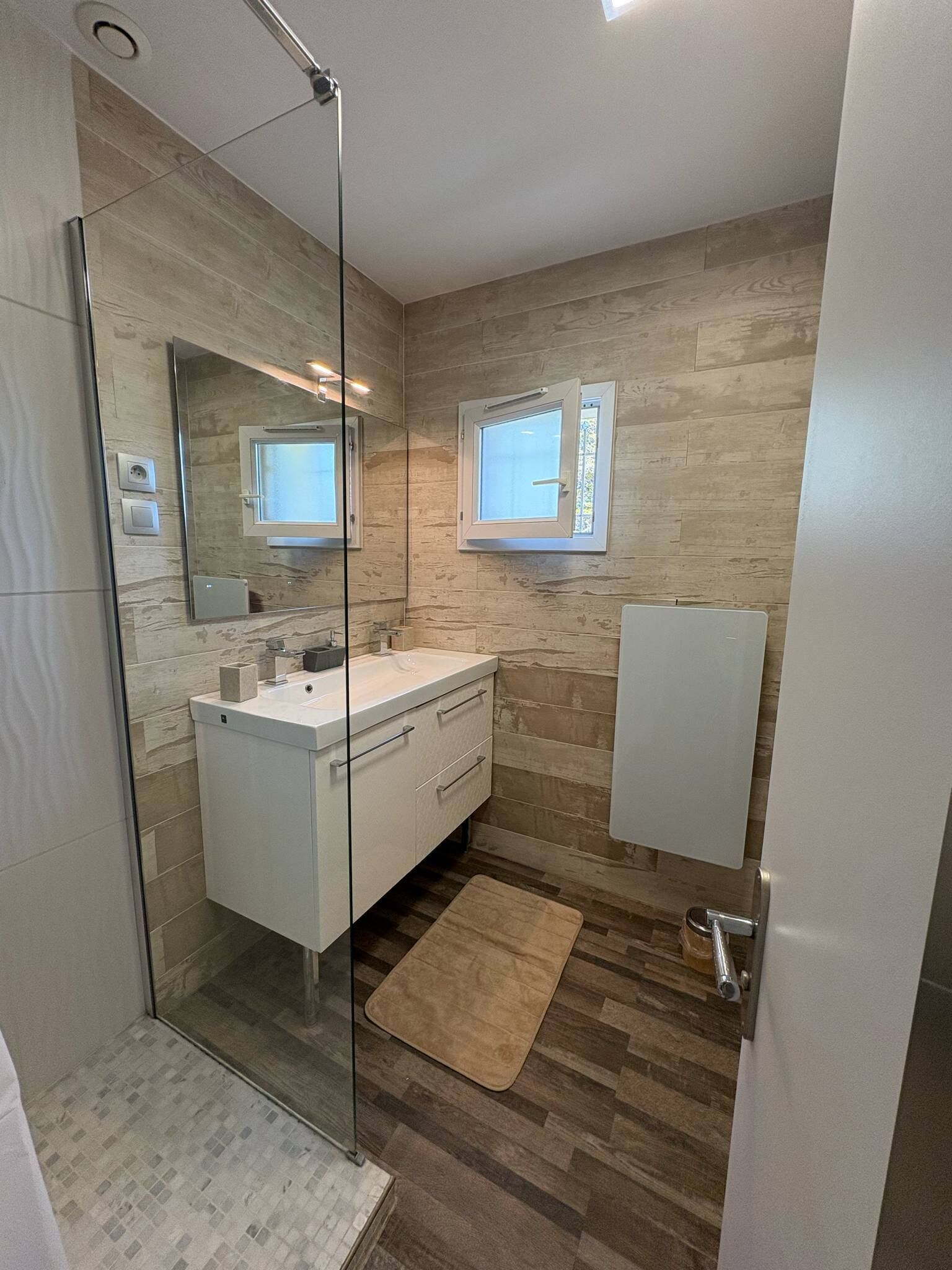 Uma casa de banho moderna com cabine de duche em vidro, toucador branco com espelho e soalho em madeira.