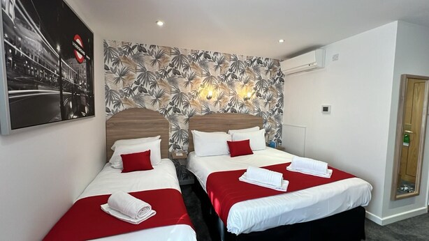 Habitación de hotel con dos camas, cada una con ropa de cama roja y almohadas blancas, papel tapiz con estampado de hojas tropicales y un cartel grande en la pared.