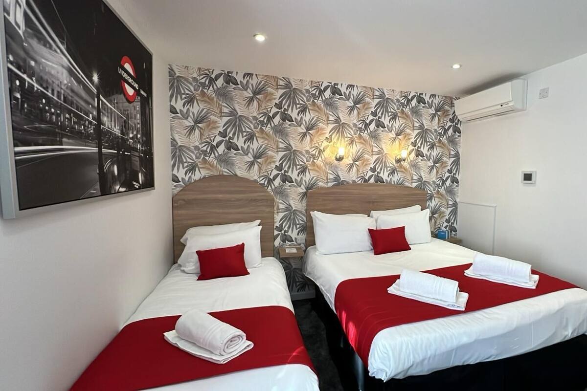 A hotel room with two beds, each with red bedding and white pillows, a tropical leaf-patterned wallpaper, and a large poster on the wall.