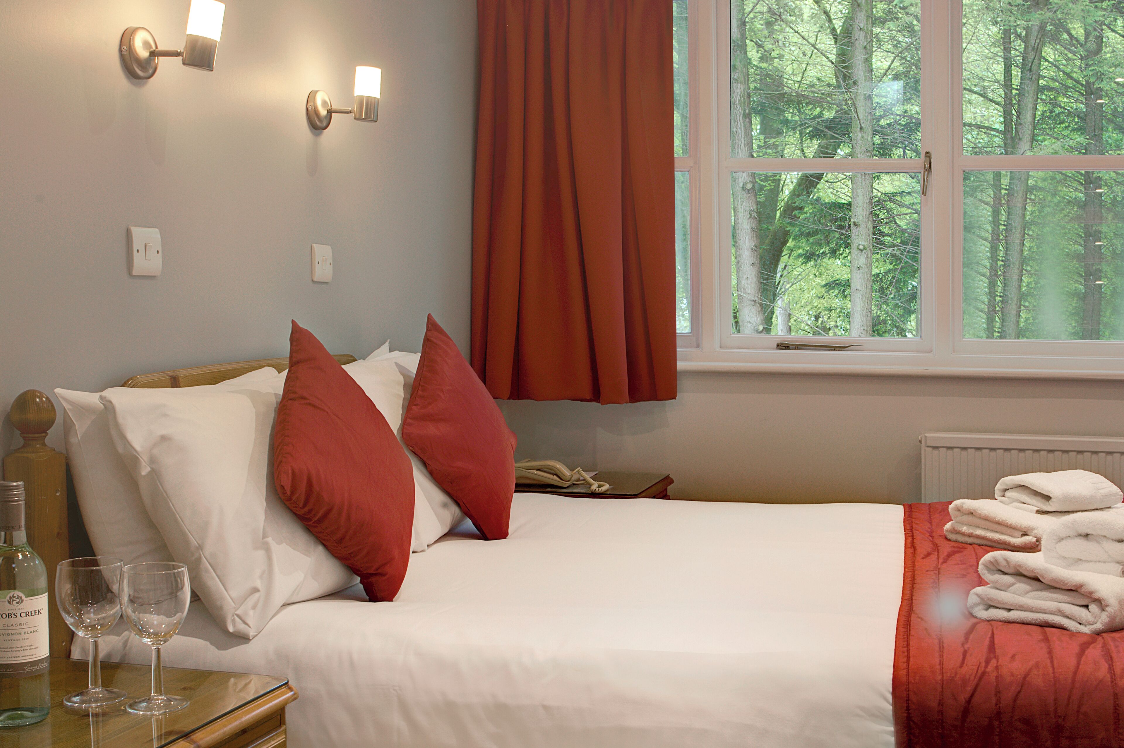 A hotel room with a bed, pillows, a window with curtains, a bedside table with a bottle and glasses, and a radiator.