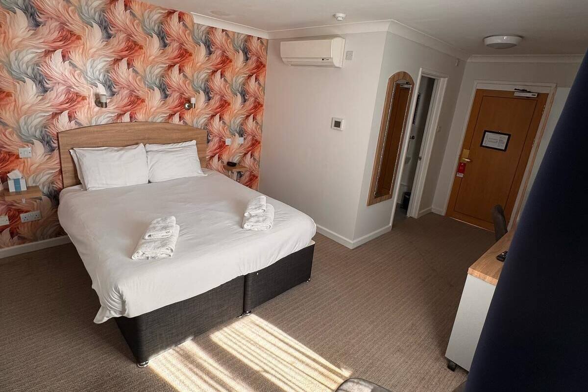 A hotel room with a bed, a desk, and a chair. There is a wall-mounted air conditioning unit and a door with a sign.