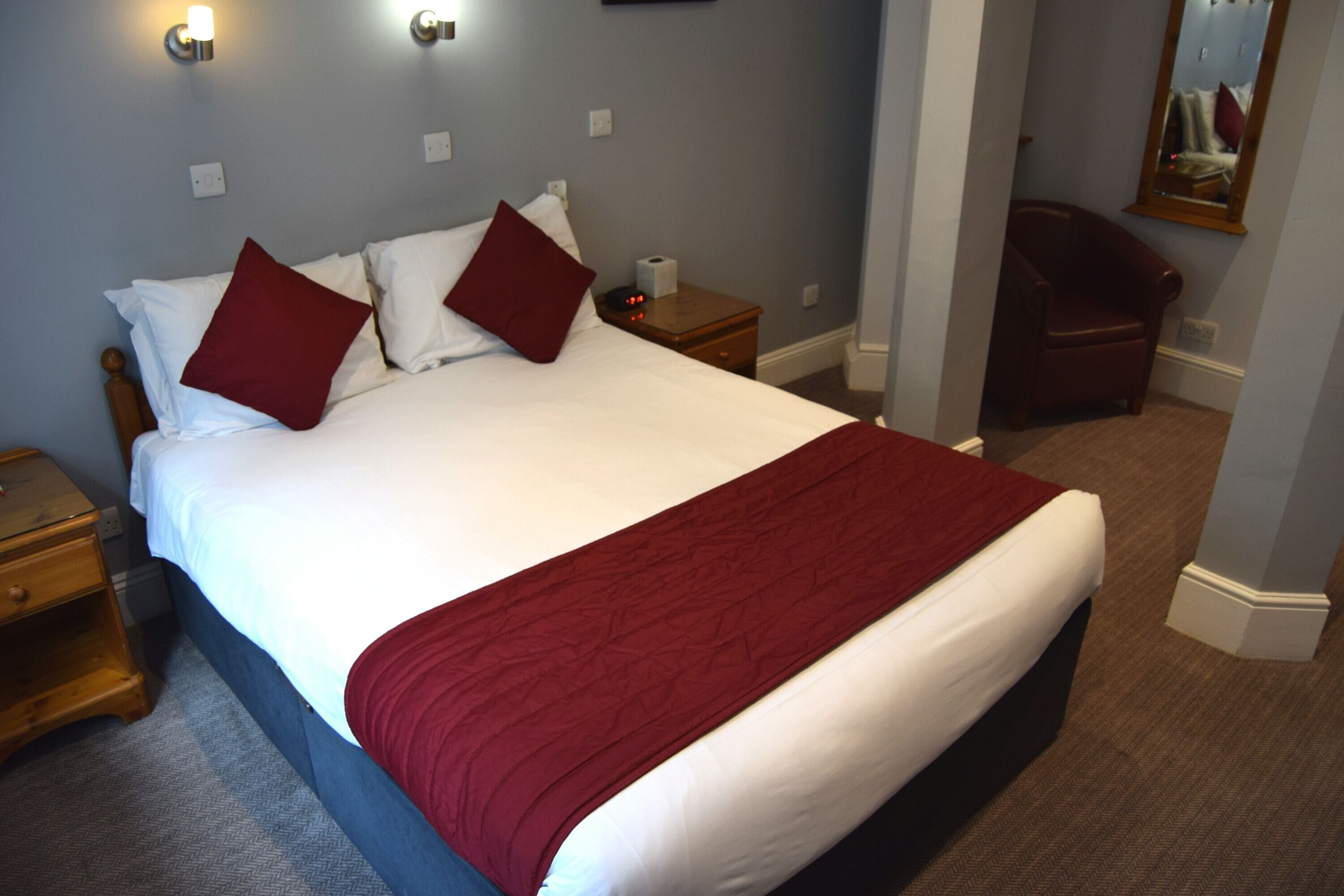 Una cama bien tendida con ropa de cama blanca y roja, una mesita de noche de madera con una lámpara, y una silla en un rincón.