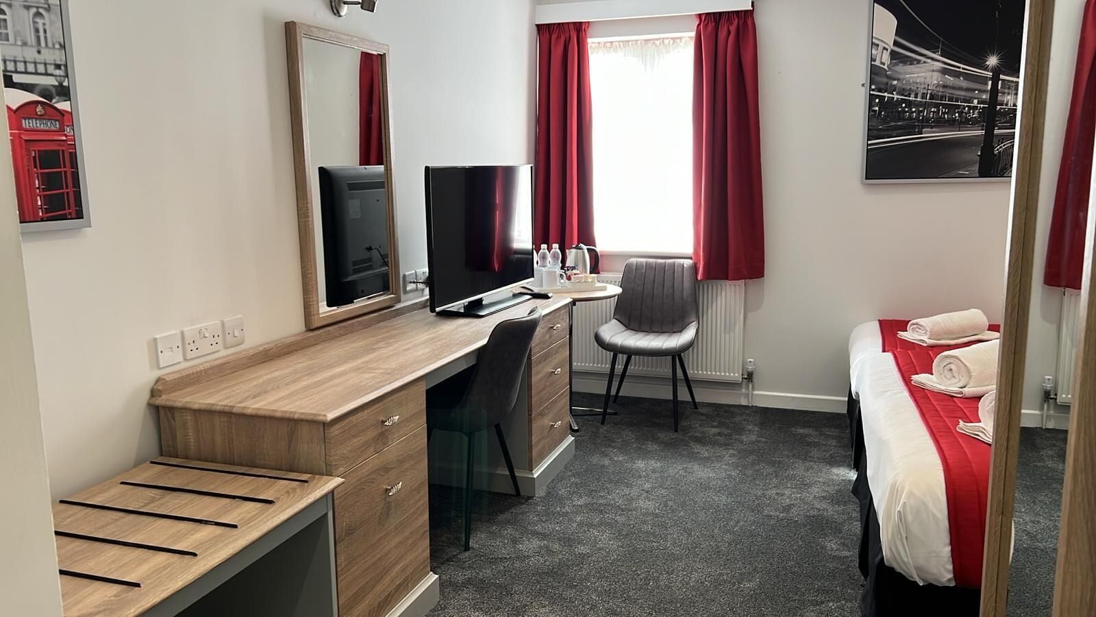 A hotel room with a wooden desk, a chair, a bed with red bedding, a mirror, and a window with red curtains.