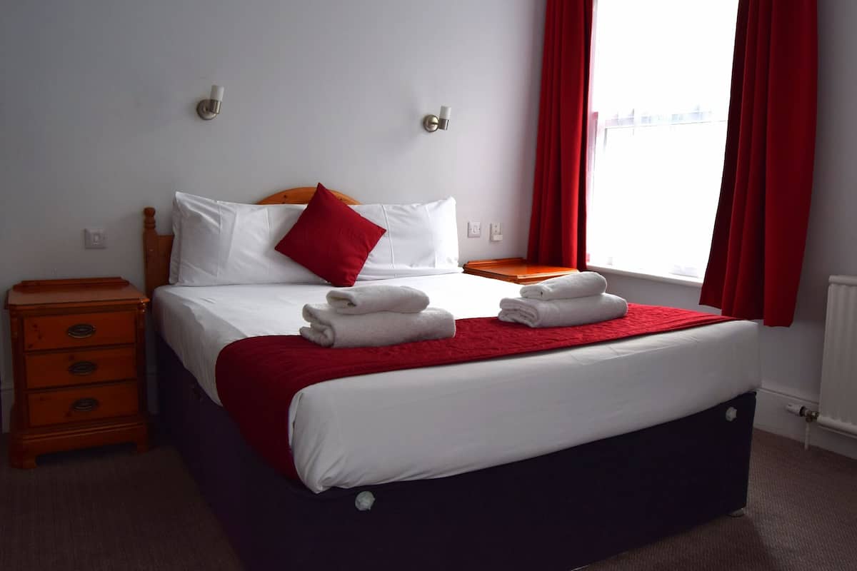 A neatly made bed with white linens and a red pillow, a wooden nightstand with drawers, a red throw blanket, and a window with red curtains.