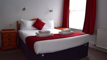 Una cama bien hecha con sábanas blancas y una almohada roja, una mesita de noche de madera con cajones, una manta roja y una ventana con cortinas rojas.