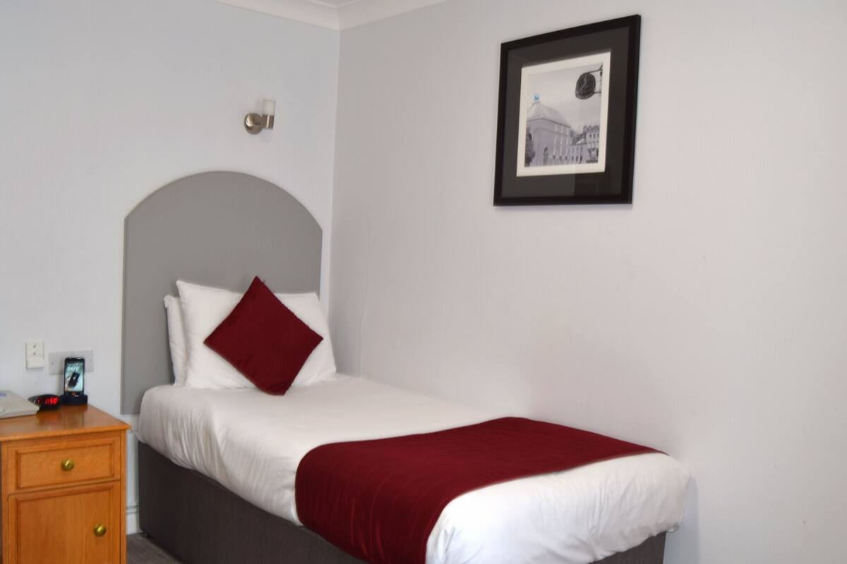 A neatly made bed with a red throw, a bedside table with a clock, and a framed picture on the wall.
