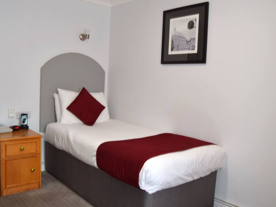 Een netjes opgemaakt bed met een rode plaid, een nachtkastje met een klok, en een ingelijste foto aan de muur.