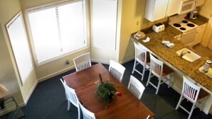 Un comedor con mesa de madera y sillas blancas, una cocina con microondas y estufa, y ventanas amplias con persianas.