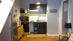 Et kompakt køkken med køleskab, mikroovn og komfur, et lille spiseområde med bord og stol, og en sofa med lampe.