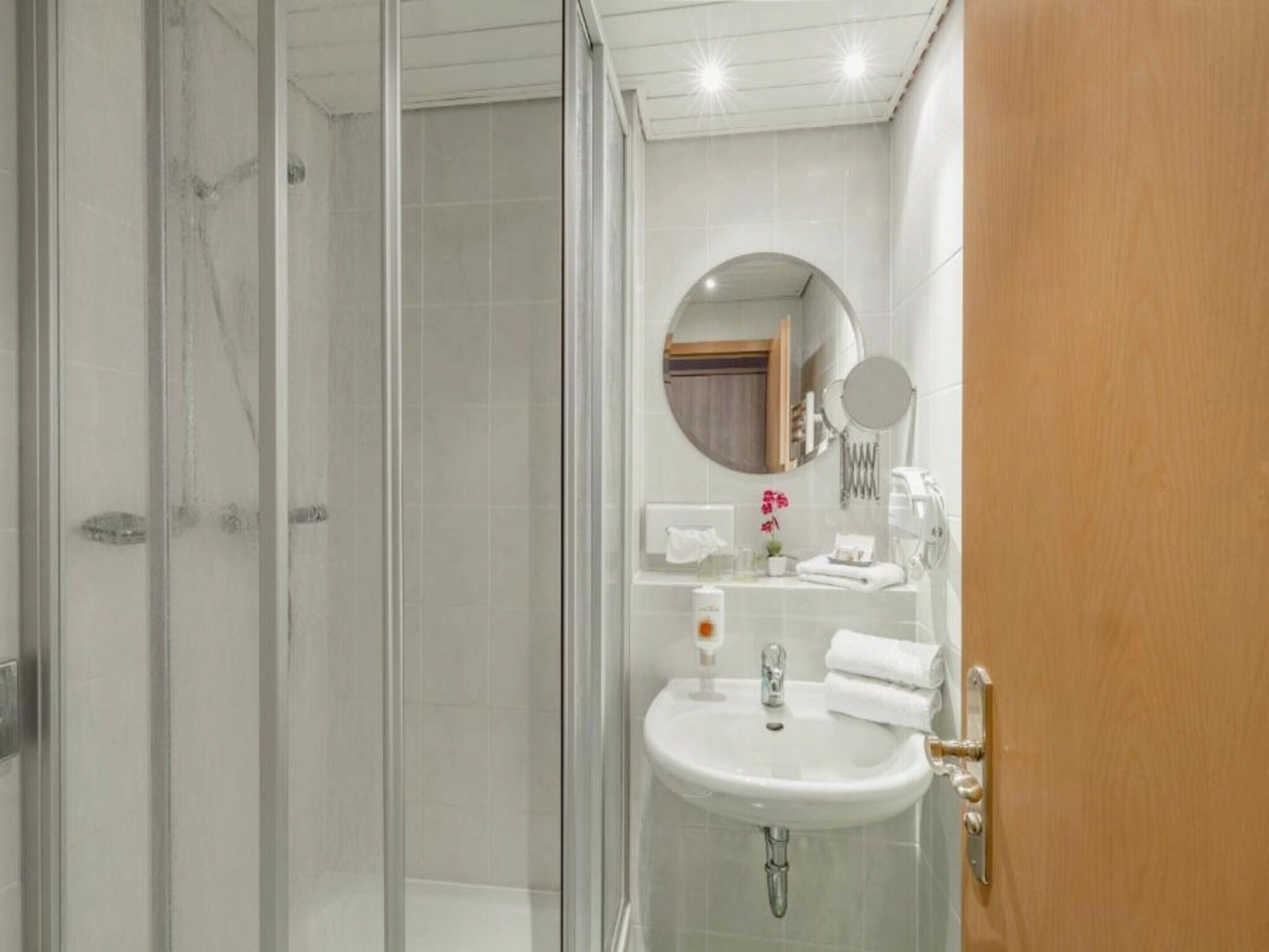 A modern bathroom with a glass shower enclosure, a white sink with a mirror above it, and a wooden door.