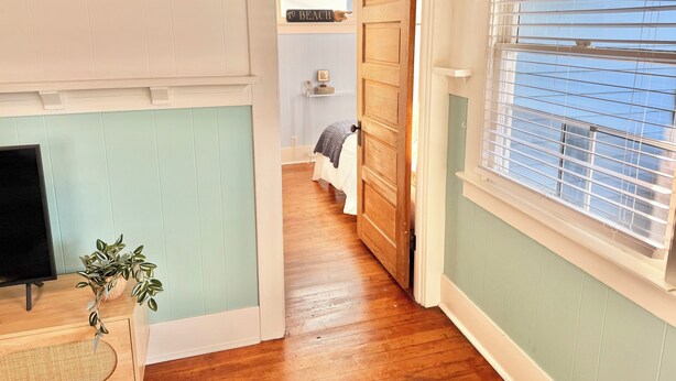 A hallway with a wooden door leading to a bedroom, a window with blinds, and a plant on a side table.