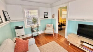 A living room with a sofa, a TV, a small table, and a chair. There is a window with blinds, a plant on a stand, and a door leading to another room.