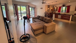 A spacious living room with a large sectional sofa, a standing fan, and a kitchen area with wooden cabinets and a modern design. - O Paradis Albion Large swimming pool and the beach just steps away (Albion)