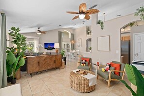 A modern living room with a wooden cabinet, a television, a ceiling fan, and a dining area in the background. - 5★ New 4BR 3BA - 3 mi to WDW, tropical Pool, Spa with all day sun, Tiki Gameroom (Four Corners)
