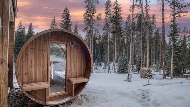 Una cabaña de madera con una puerta circular, un banco en el interior y un bosque nevado de fondo.