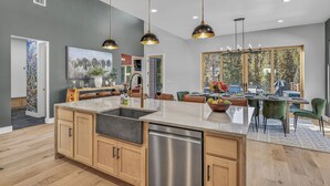 Cocina moderna con una isla central, armarios de madera, electrodomésticos de acero inoxidable, iluminación colgante y vistas al exterior a través de amplios ventanales.