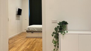 Moderni huone, jossa on puulattia, valkoinen vaatekaappi, yöpöytä, jossa on kasvi, ja seinälle kiinnitetty kello.
