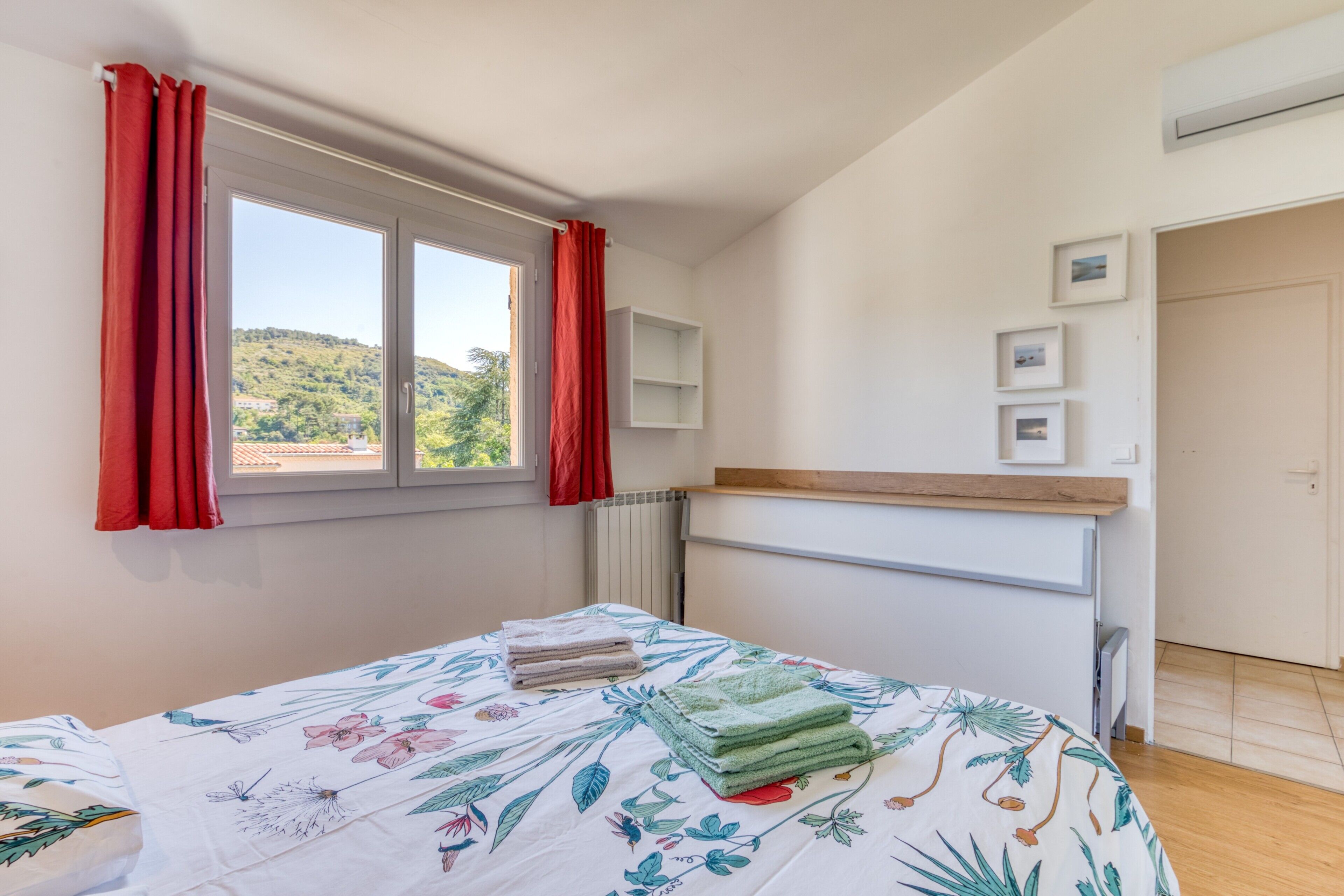 A bedroom with a bed, a window with red curtains, a radiator, and a wall-mounted shelf with pictures.