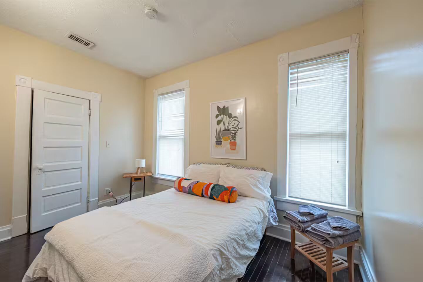 Une chambre à coucher avec un lit, une table de chevet, une plante fixée au mur et une fenêtre avec des stores.