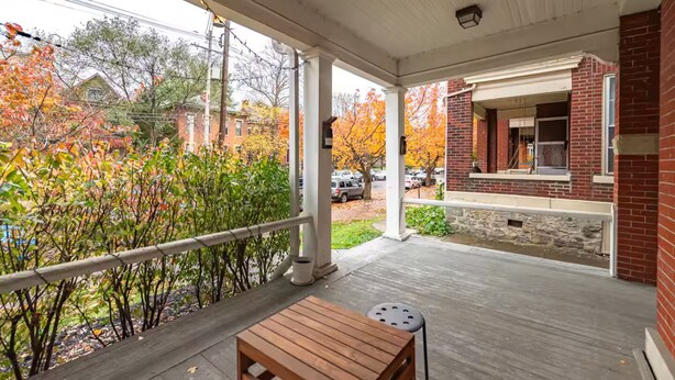 A covered porch with a wooden table and a metal stool, overlooking a street with autumn foliage and brick buildings. - 3-Bedroom in Downtown Columbus (Columbus)