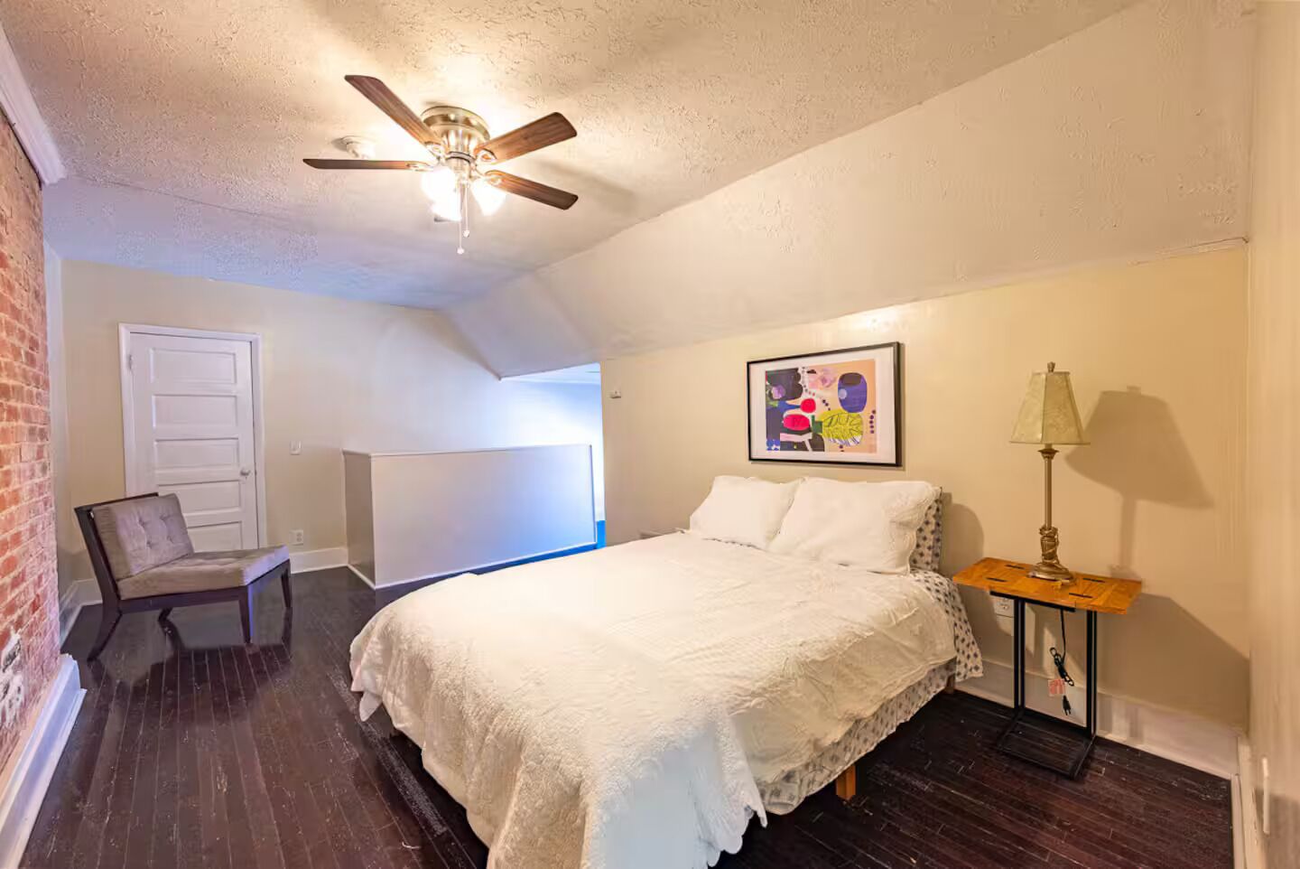 A bedroom with a bed, a ceiling fan, a chair, a lamp, and a painting on the wall.