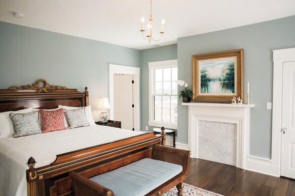 A bedroom with a large bed, a bench, a fireplace, and a painting on the wall.