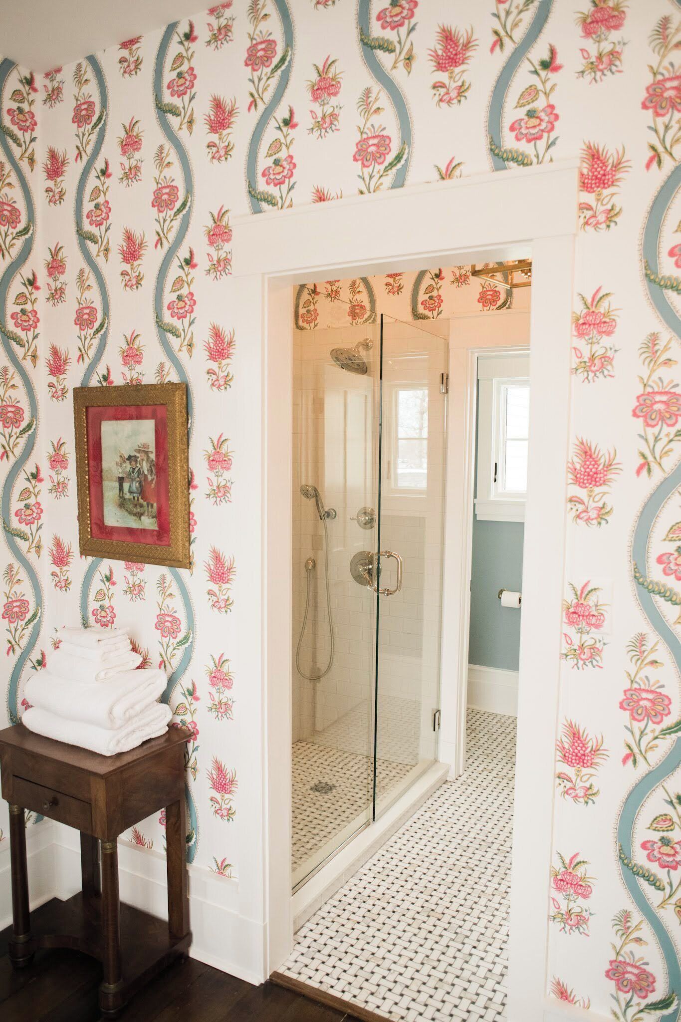 Une salle de bain avec un papier peint à motifs floraux, une porte de douche en verre et un petit tabouret en bois sur lequel sont posées des serviettes.