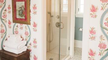 Une salle de bain avec un papier peint à motifs floraux, une porte de douche en verre et un petit tabouret en bois sur lequel sont posées des serviettes.