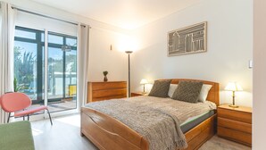 Un dormitori amb llit, taules de nit, una cadira i vista a través d'una porta de vidre.