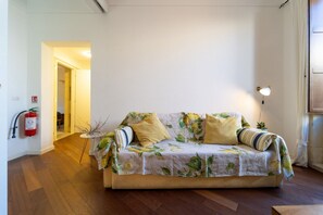 A room with a floral-patterned sofa, a small round table, and a wall-mounted lamp. - GuestHost - Cozy and bright 87 square meters apartment, perfect for five people, located on the second floor of a building (no elevator).The property is located on the island of Ortigia (Restricted-Traffic Area), the historic and cultural heart of Syracu (Syracuse)