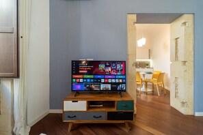 Ein Wohnzimmer mit einem Flachbildfernseher auf einem Holzständer, ein Essbereich mit Tisch und Stühlen sowie eine durch eine offene Tür sichtbare Küche.