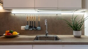 Una cocina con un lavamanos de acero inoxidable, un cuenco de fruta, una planta en maceta y utensilios colgantes.