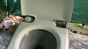 Un baño químico con asiento elevado y un recipiente central, ubicado al aire libre con un fondo de malla verde.
