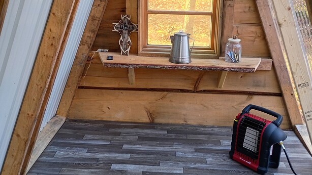 Un dormitorio estilo altillo de madera con techo inclinado, una ventana, un hervidor de metal, un frasco de vidrio y un calentador portátil en el suelo.