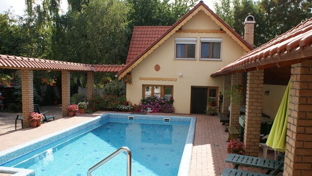 En swimmingpool med ett kaklat poolområde, ett hus byggt av tegel med ett rött tak och utomhusmöbler.