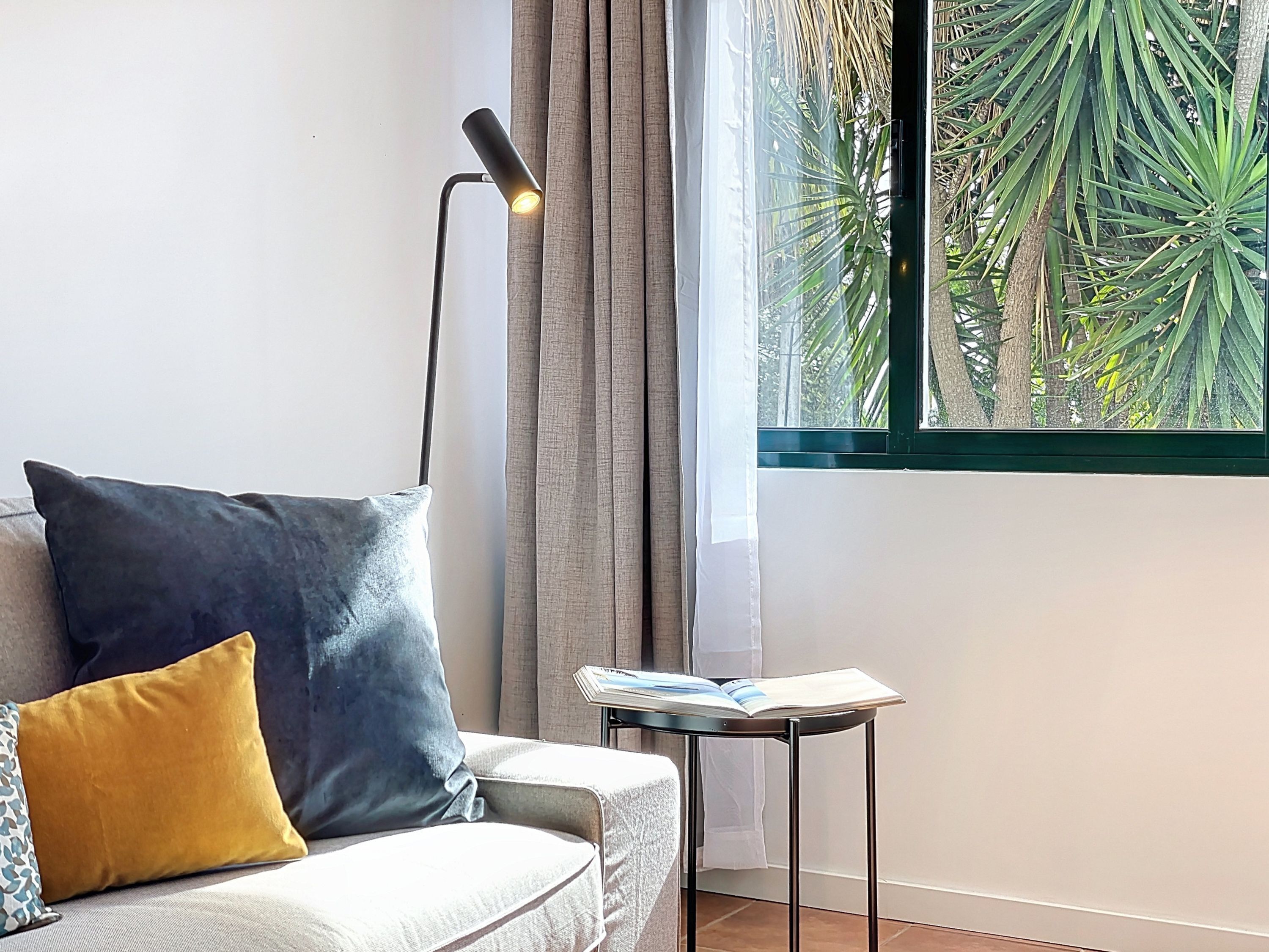 Ein modernes Wohnzimmer mit einer Couch, einem Beistelltisch mit einem Buch, einer Stehlampe und einem Fenster mit Blick auf Grünflächen.