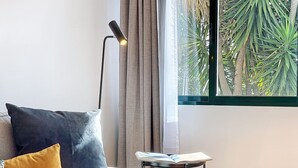 Ein modernes Wohnzimmer mit einer Couch, einem Beistelltisch mit einem Buch, einer Stehlampe und einem Fenster mit Blick auf Grünflächen.