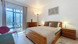 Ein Schlafzimmer mit einem hölzernen Bett, zwei Nachttischen mit Lampen, einem roten Sessel und Blick auf einen Balkon mit Gartenmöbeln.
